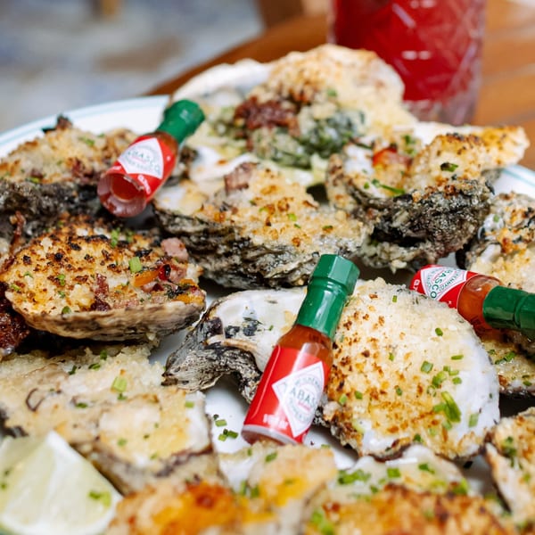 A plate of baked oysters topped with cheese, bacon, and chives, with mini Tabasco bottles.