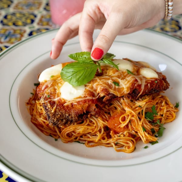 A hand garnishes a plate of chicken parmesan with spaghetti and fresh basil.