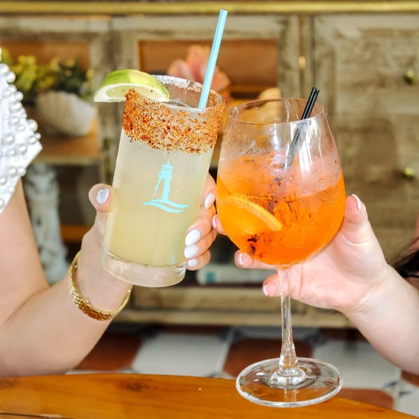 Two people clinking glasses, one with a Lighthouse Coastal Brasserie margarita, the other with an Aperol Spritz.