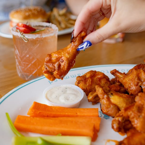 Hand holding a buffalo wing over a plate of wings, dip, carrots, and a cocktail.