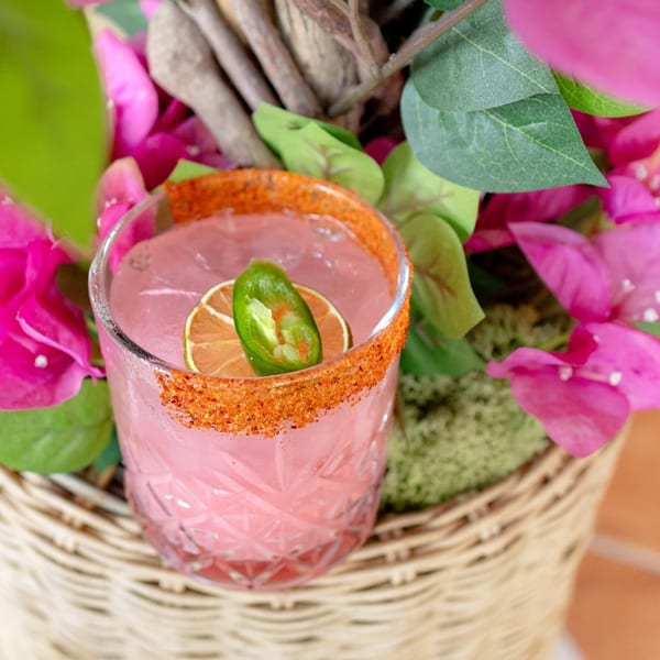 Pink cocktail with a chili-lime rim, lime slice, and jalapeño, nestled in a wicker basket with pink flowers.